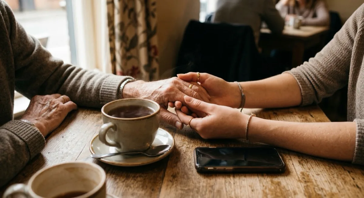 An elderly person and younger family member holding hands across a table with tea